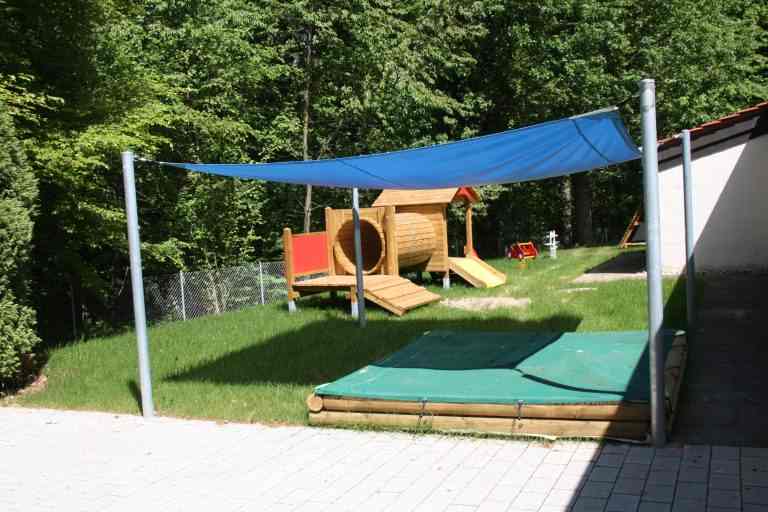 Sandkasten aus Holz auf einem Spielplatz, abgedeckt mit grüner Netzplane und darüber gespanntes, blaues Sonnensegel.
